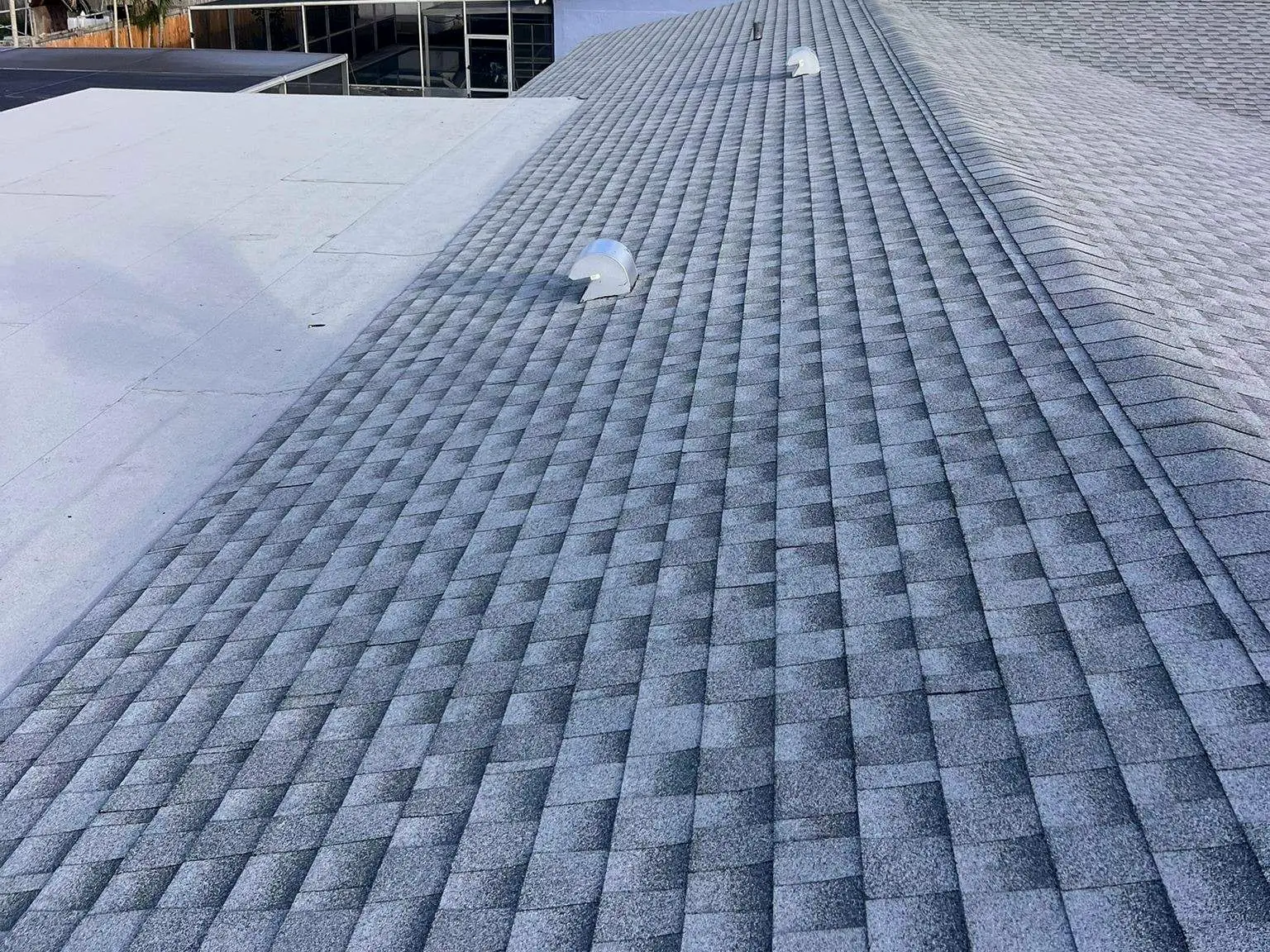 Finished shingle roof with roof vents along the slope, beside a flat roof section